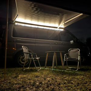 LED awning lighting on VW camper van at night with outdoor camping chairs and table