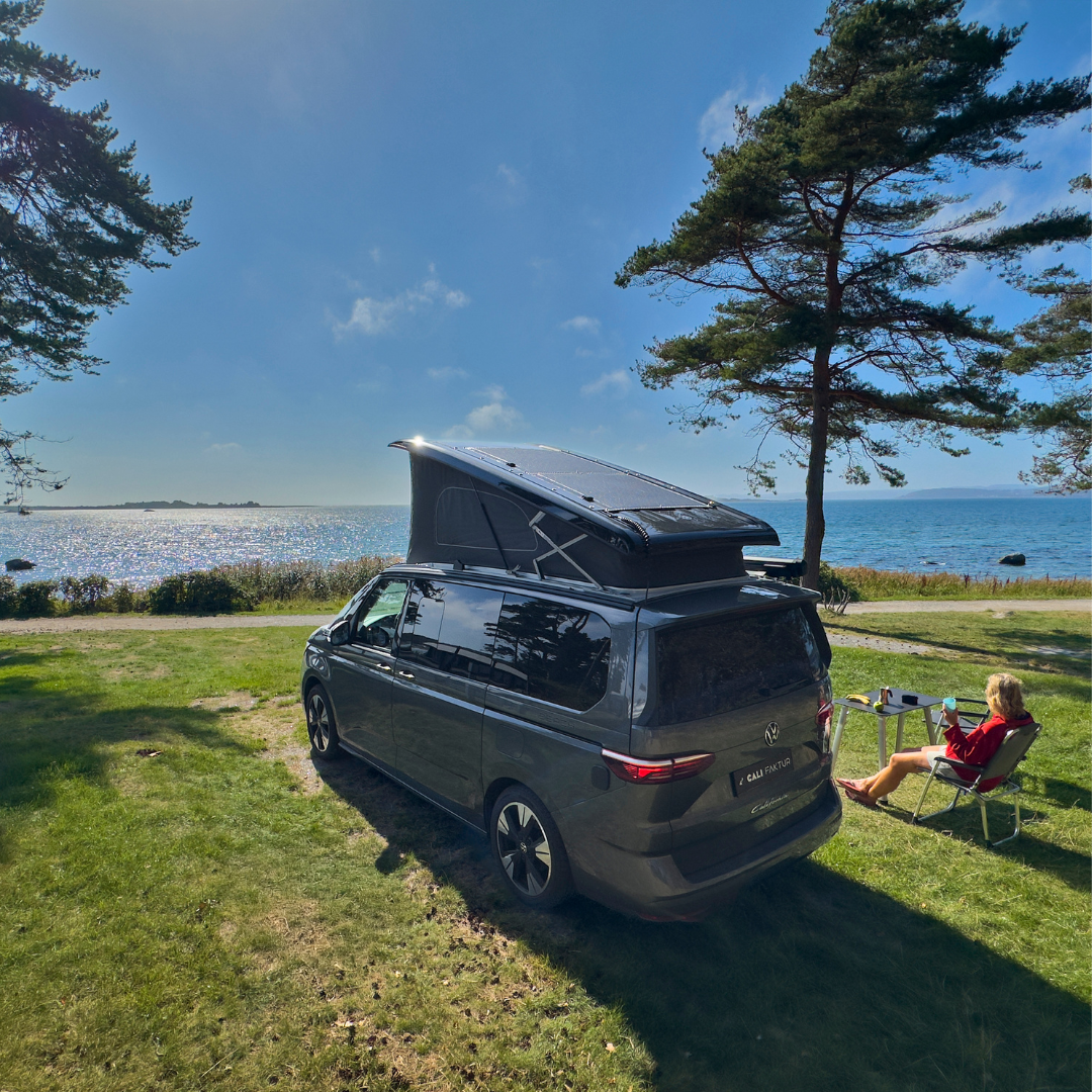 VW California T7 mit CALIFAKTUR Solarmodul am Meer, Frau entspannt mit Getränk im Campingstuhl in der Sonne